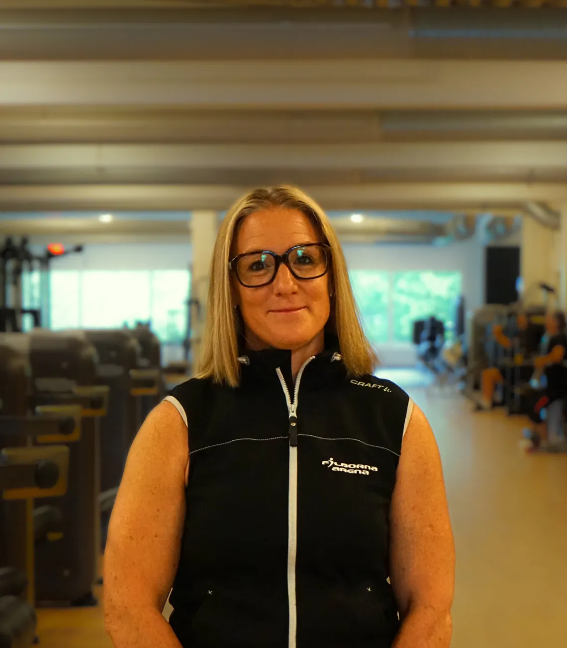 En person som bär glasögon och en ärmlös svart jacka står framåtvänd i en gymmiljö med träningsredskap och människor i bakgrunden. Jackans text lyder "Filborna Arena" och "CRAFT."