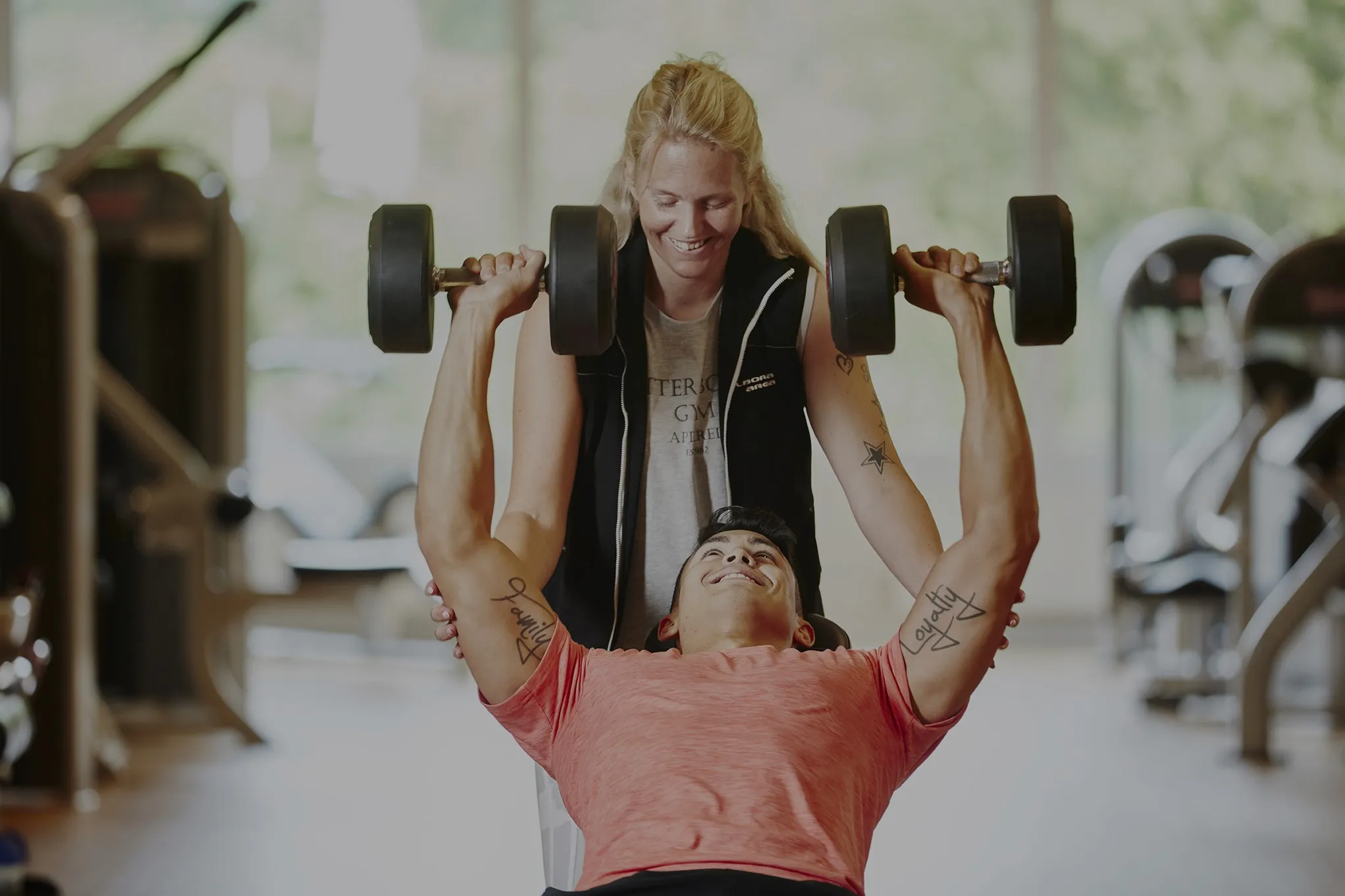 En person i en röd tröja lyfter hantlar, assisterad av en leende tränare på ett gym. Tatuerade armar är synliga, med gymutrustning i den suddiga bakgrunden.