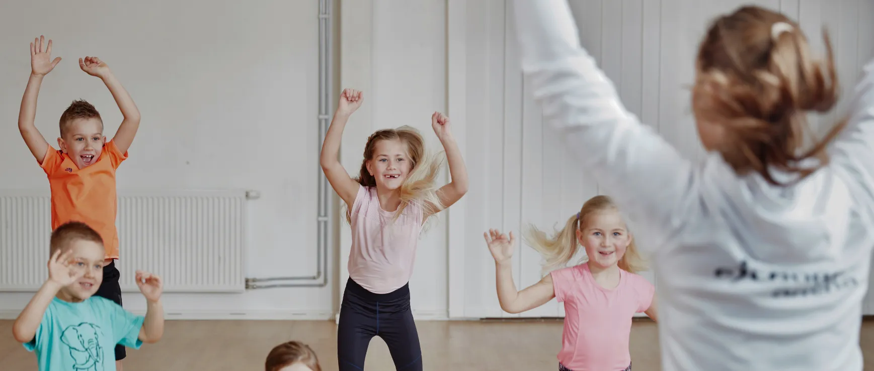 Barn höjer med glädje sina armar och deltar i en energisk aktivitet ledd av en instruktör i ett ljust inomhusutrymme med vita väggar och ett trägolv.